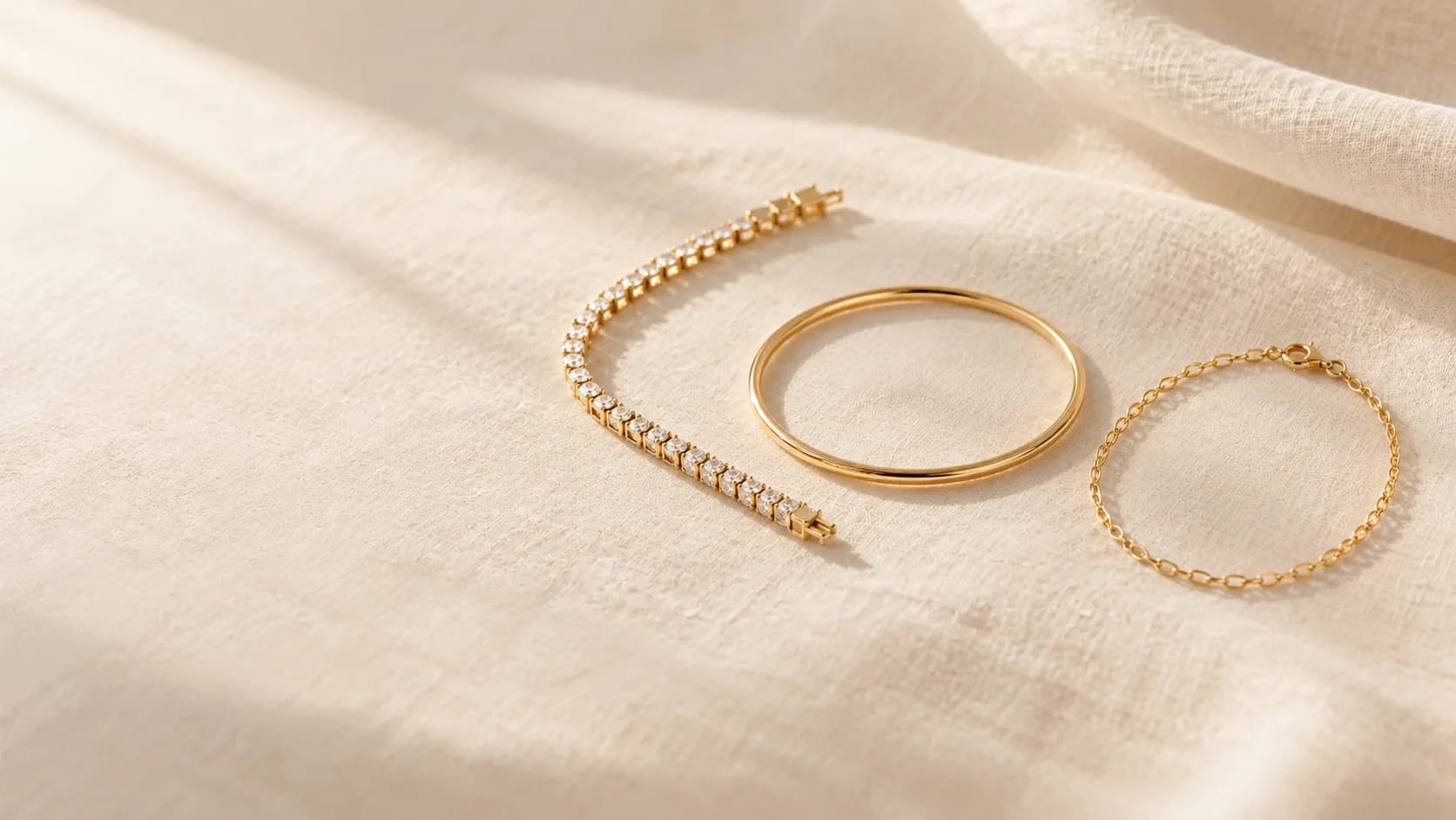 a diamond tennis bracelet, a gold bangle and a fine gold chain bracelet laid on cream linen